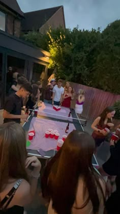 a group of people standing around a ping pong table at a party with drinks on it