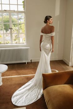 a woman in a white wedding dress is standing near a couch and looking out the window