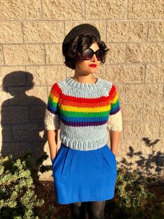 a woman standing in front of a brick wall wearing a rainbow sweater and blue skirt