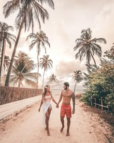 a man and woman holding hands walking down a dirt road with palm trees in the background