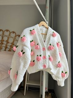 a white sweater with pink roses on it hanging from a clothes rack next to a bed