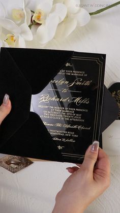 a person holding up a black and gold wedding card with white flowers in the background