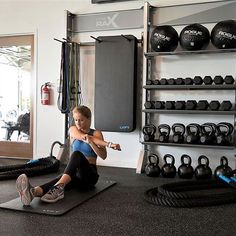 a woman sitting on a mat in a gym with kettles and exercise mats behind her