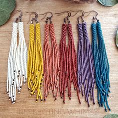 six different colored seed beaded earrings hanging from hooks on a wooden table next to leaves