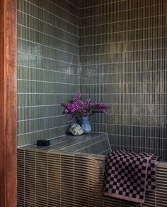 a vase with purple flowers sitting on top of a wooden shelf next to a tiled wall