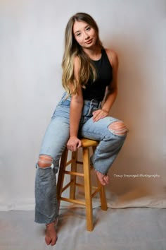 a woman sitting on top of a wooden stool wearing ripped jeans and black tank top