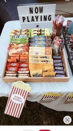 an outdoor movie party with popcorn, snacks and candy bar sign on the table that says now playing