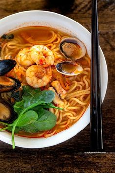 a white bowl filled with noodles and seafood next to chopsticks on a wooden table