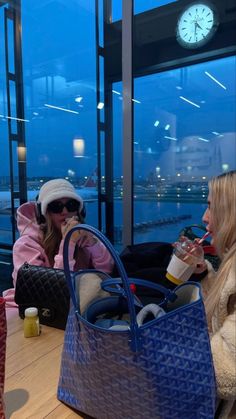 two women sitting at a table with drinks in front of a large window looking out onto the water