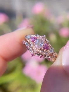 a person holding a ring with pink and white stones on it's side in front of some flowers