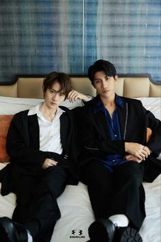 two young men sitting on top of a bed next to each other in black suits