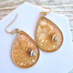 "These exquisite Boho Handmade Coral Peach Translucent Resin Seashell Starfish Ocean Themed Sealife Teardrop Dangle Earrings are perfect for adding a touch of seaside charm to your jewelry collection. Each earring features a beautifully crafted translucent resin teardrop in a refreshing mint color, reminiscent of the ocean's tranquil hues. Encapsulated within the resin are delicate seashells and a whimsical starfish, capturing the essence of the beach and its mesmerizing sealife. Measuring at a comfortable 1.75\" (4.45 cm) drop, these earrings offer an elegant and eye-catching length, allowing them to sway gently with your movements. The teardrop shape adds a graceful touch, enhancing their overall appeal. These earrings also make fantastic vacation gifts, serving as a delightful reminder Gold Starfish Shell For Beach, Summer Beach Teardrop Earrings, Gold Dangle Teardrop Earrings For Beach, Gold Teardrop Earrings For Summer, Beach Dangle Teardrop Earrings With Ear Wire, Handmade Teardrop Earrings For Beach, Black Gold Necklace, Boho Handmade, Teardrop Dangle Earrings