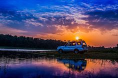 an suv is parked on the bank of a body of water as the sun sets