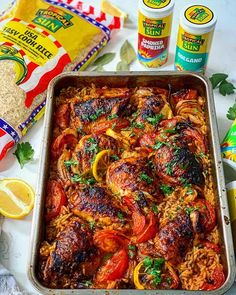 a pan filled with chicken and tomatoes next to some seasoning, lemon wedges and garlic