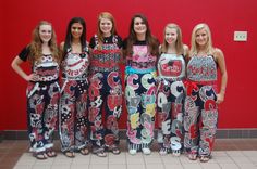 four girls standing in front of a red wall wearing different colored pants and dresses with designs on them