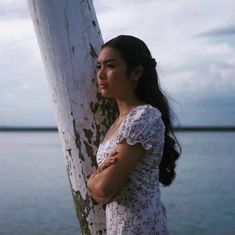 a woman standing next to a tree near the water with her arms folded on it