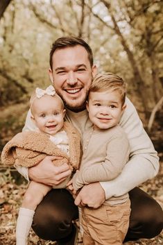 a man holding two small children in his arms and smiling at the camera with trees behind him