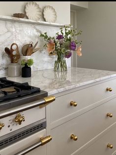 a white kitchen with marble counter tops and gold pulls on the stove top, along with flowers in a vase