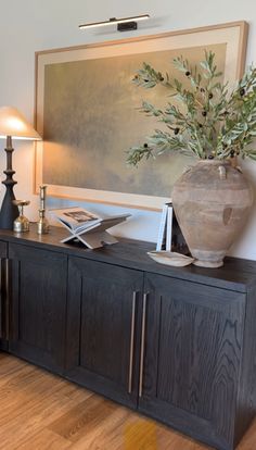 a vase with some flowers on top of a wooden cabinet next to a lamp and pictures