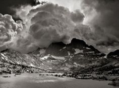 black and white photograph of mountains with clouds in the sky above them, near a body of water