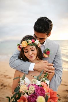 a bride hugging her groom on the beach with flowers in his hair and wearing a flower crown