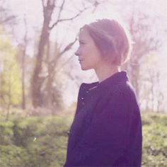 a woman standing in the middle of a forest looking off into the distance with trees and bushes behind her