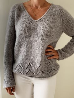 a woman wearing a gray sweater and white pants with her hands on her hips, standing in front of a wall