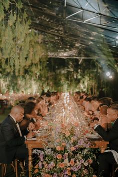 a group of people sitting around a long table with flowers on it and greenery hanging from the ceiling