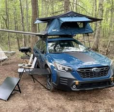 a blue subarunt parked in the woods next to a camp table and chair