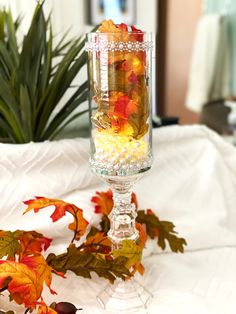 a glass vase filled with autumn leaves on top of a white table cloth next to a potted plant