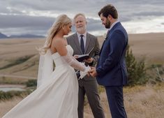 a bride and groom holding hands during their wedding ceremony in an open field with mountains in the background