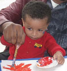 a young boy sitting in front of a painting with red paint on it and holding a knife