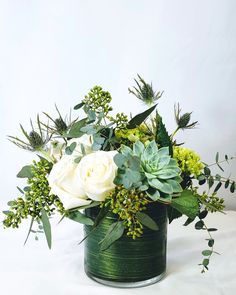 a green vase with white flowers and greenery in it on a white tablecloth