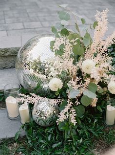 a vase with flowers and candles sitting on the ground in front of some bushes next to a metal ball