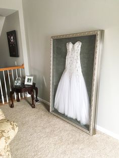 a wedding dress hanging in a frame next to a chair and table with pictures on it