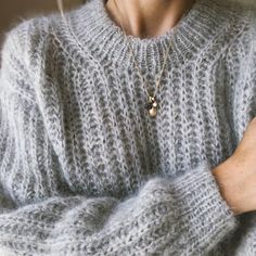 a woman wearing a gray sweater with a gold necklace on her neck and arms crossed