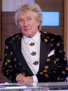 an older man in a black and gold suit sitting down with his hands on his hips