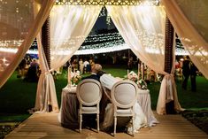 two people sitting at a table with white chairs under a canopy covered in fairy lights