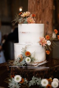 a three tiered white cake with orange and white flowers on the top is surrounded by greenery