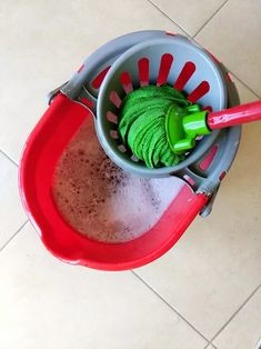 a red and gray bucket filled with green stuff on top of a tiled floor next to a red plastic mop