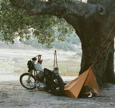 a motorcycle parked next to a tree with a tent on it's back ground