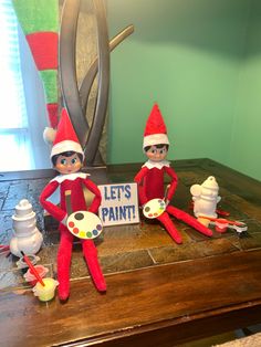 two elfs sitting on top of a table with paints and paintbrushes in front of them