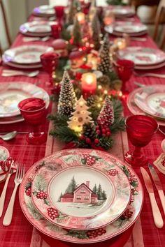 a christmas table setting with red and white plates, silverware, candles and pine trees