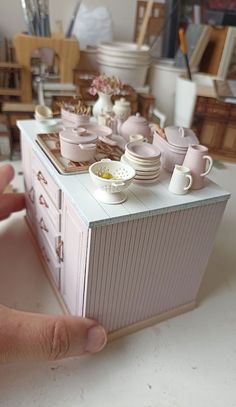 a hand is holding a dollhouse table with cups and saucers on it,