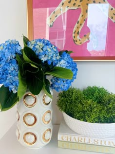 blue flowers in a white vase next to a green plant and a leopard painting on the wall