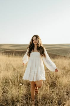 a woman in a white dress is walking through the tall grass with her hands behind her back