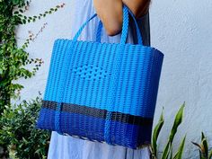 a woman holding a blue woven bag in front of a white wall and green plants