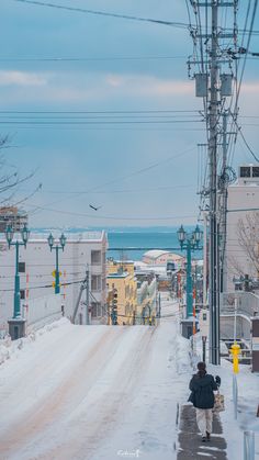 a woman walking down a snow covered street next to power lines and telephone poles with the ocean in the background