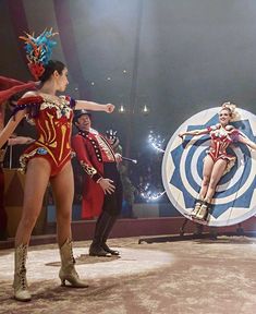 two women in costumes are performing on stage with other performers behind them and one is holding a shield