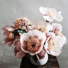 a white vase filled with pink flowers on top of a wooden table next to a wall